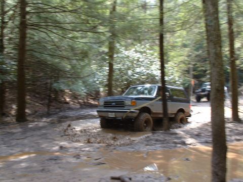 1991 Ford Bronco - Ford Bronco navigates a muddy path in a wooded area, with a second vehicle following behind.