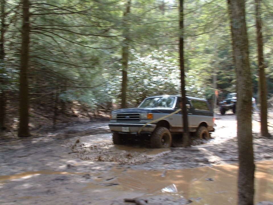 1991 Ford Bronco driving through a muddy trail in a forest, another vehicle visible in the background