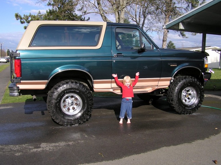 Green 1995 Ford Bronco with tan trim, lifted suspension, and large tires, a child stands in front of it