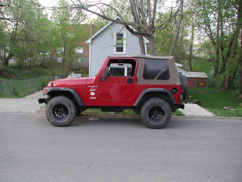 1998 Jeep TJ - A red Jeep Wrangler is parked on a residential street with a house and trees in the background.
