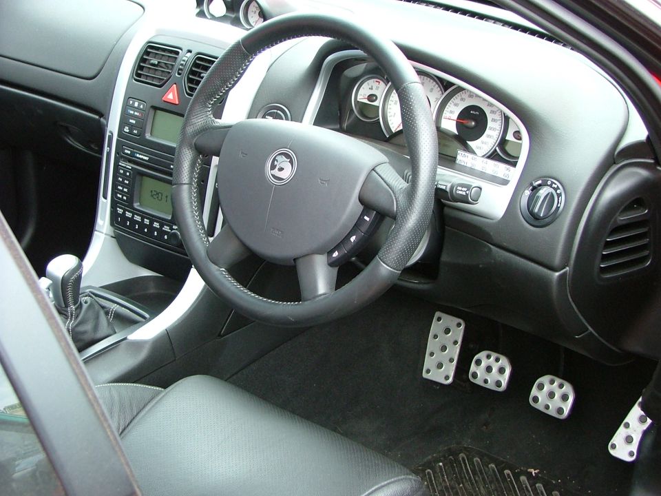 Car interior with leather seats, steering wheel, dashboard, and pedals. Watermark "SUPERMOTORS" visible