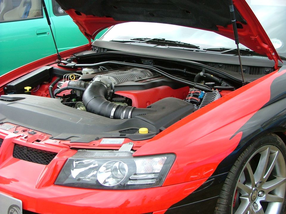 Engine bay with supercharger and red accents. A green car is visible to the left