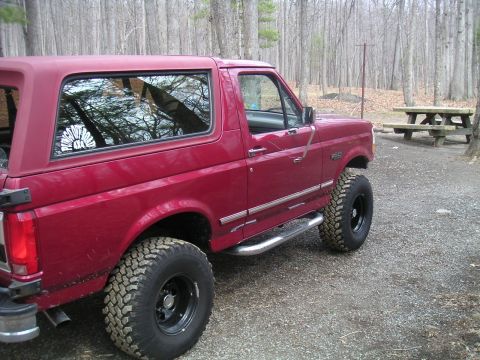 1994 Ford Bronco - A red Ford Bronco with off-road tires sits parked in a wooded area next to a picnic table.