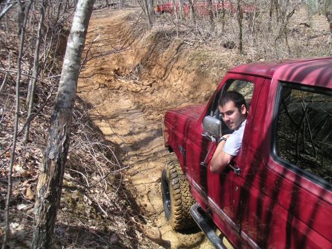 1994 Ford Bronco - Man driving a red Ford Bronco on a muddy, wooded trail. Another red vehicle is visible in the background.