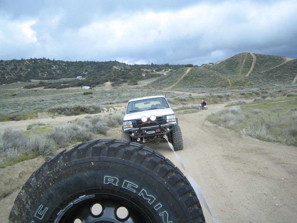 White pickup truck with winch and lights, towing another vehicle on a dirt road. Tire in foreground