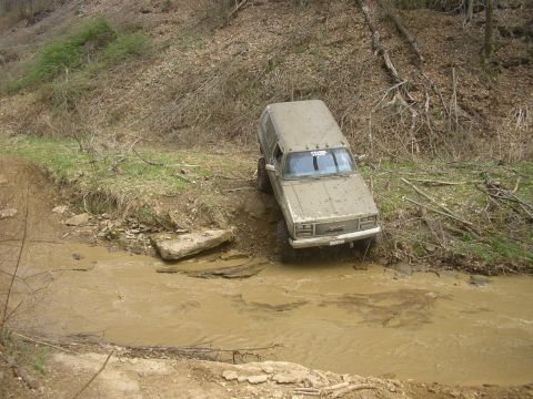1989 GMC Jimmy - This GMC SUV is navigating a muddy creek on an off-road trail, with trees and debris on the surrounding slopes.