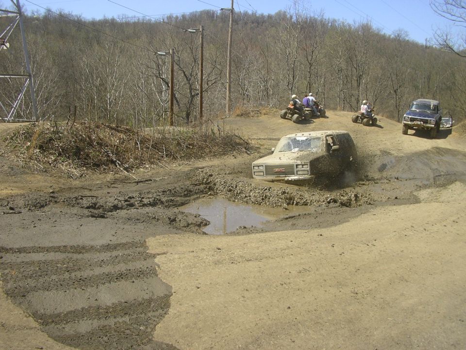 Muddy GMC SUV driving through a large mud puddle, with ATVs and another truck in the background