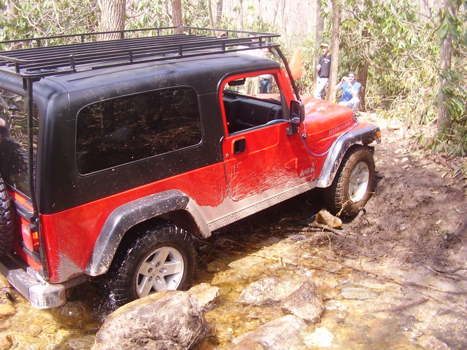Red Jeep Wrangler with black hardtop and roof rack driving through a rocky, muddy stream