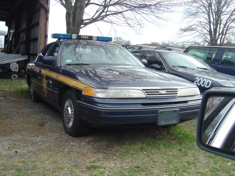 2001 Ford Crown Victoria - Police car with light bar and markings, parked outdoors next to other vehicles.