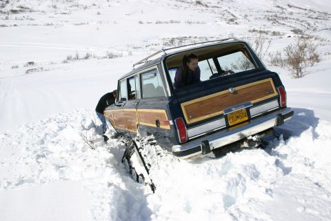 2003 Jeep TJ - A dark blue Jeep Grand Wagoneer with wood paneling is stuck in deep snow, with people attempting to free it.