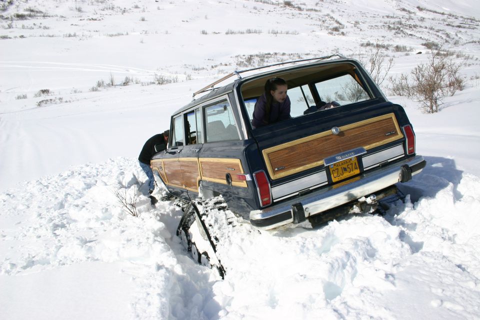 Blue woodgrain Jeep Grand Wagoneer stuck in deep snow with Alaska plate, rear three-quarter view
