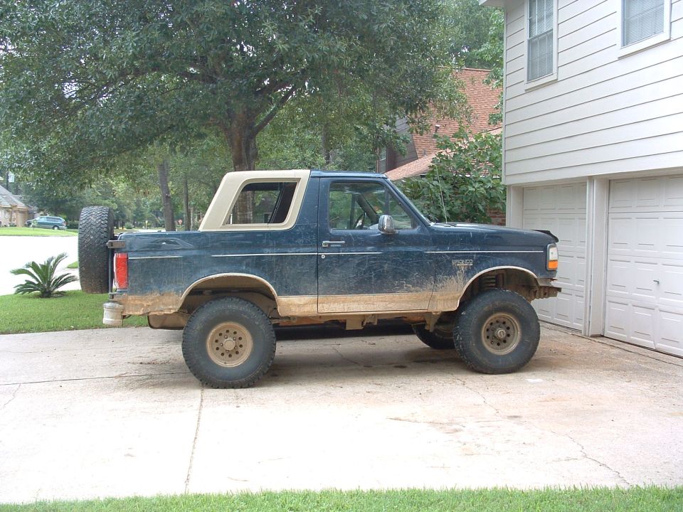Muddy dark blue Ford Bronco with top removed, lifted on mud tires in a driveway
