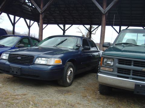 2001 Ford Crown Victoria - A blue 2001 Ford Crown Victoria is parked next to a green pickup truck under a covered structure.