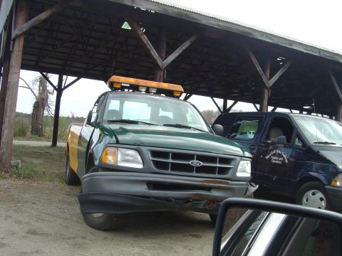 2001 Ford Crown Victoria - Green Ford F-150 pickup truck with a light bar and damaged bumper is parked next to a dark blue van.