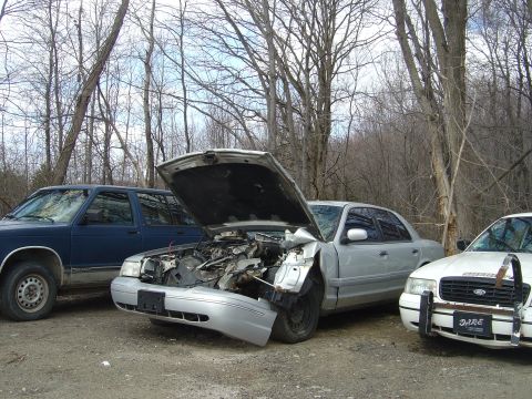 2001 Ford Crown Victoria - Three vehicles are parked outdoors in a wooded area, one silver sedan has significant front-end damage.