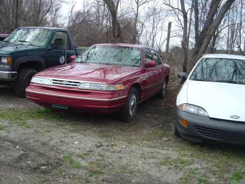 2001 Ford Crown Victoria - A red 2001 Ford Crown Victoria sedan is parked outdoors between a green pickup truck and a white car.