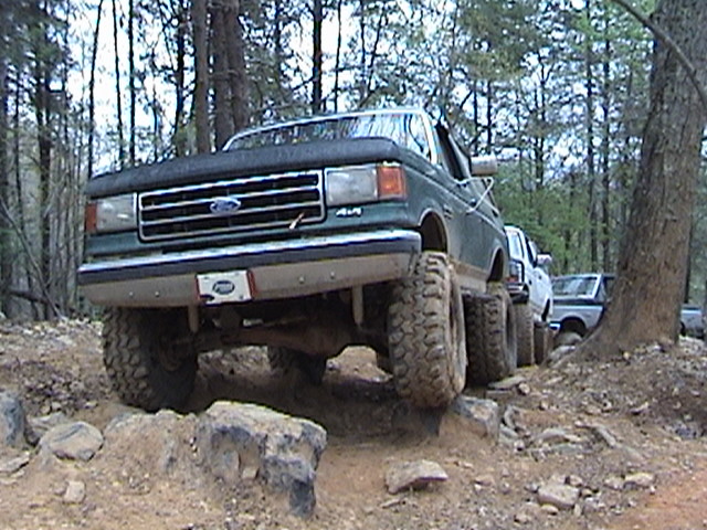 Green Ford F-Series truck with lifted suspension and large tires driving on a rocky trail. Other trucks are behind it
