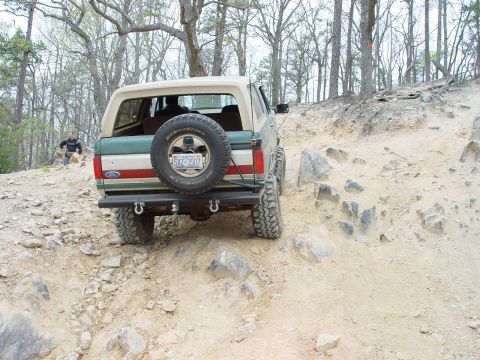 1996 Ford Bronco - A Ford Bronco navigates a challenging off-road trail with trees in the background.