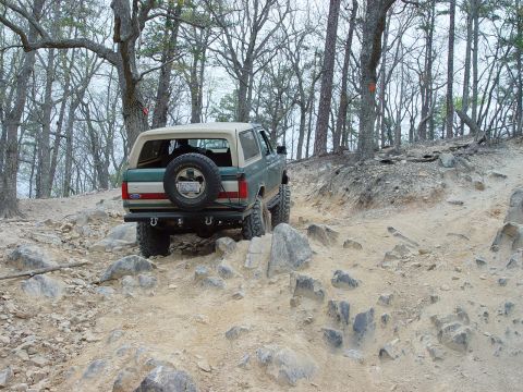 1996 Ford Bronco - A 1996 Ford Bronco navigates a challenging, rocky off-road path surrounded by trees.