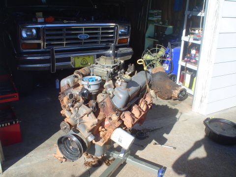 1984 Ford Bronco - Engine removed from a vehicle and placed on a stand, with the front of a Ford truck visible in the background.