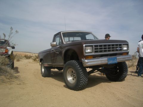 1985 Ford Ranger - A lifted brown pickup truck sits on a dusty desert road next to another vehicle.