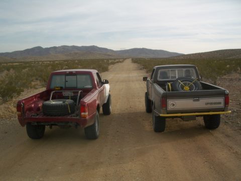 1985 Ford Ranger - Two pickup trucks drive down a dusty desert road with mountains in the background.