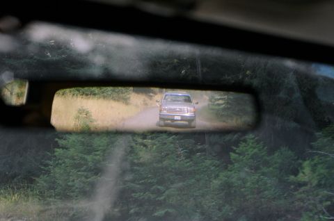 1979 Ford Bronco - A silver pickup truck drives down a dirt road surrounded by trees, as seen through a rearview mirror.