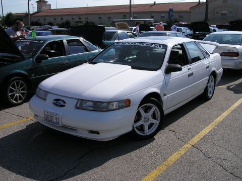 1993 Ford Taurus - A white sedan is parked in a lot with other vehicles, some with hoods open.