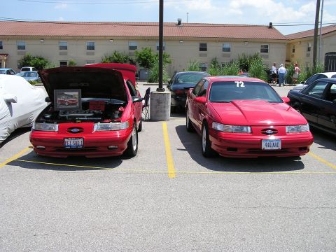 1993 Ford Taurus - Two red Ford Taurus sedans are parked in a lot, one with its hood open, showing a framed picture inside.