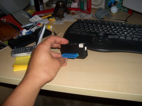 1990 Ford Bronco - A hand holds a black electronic device with a blue connector, resting on a desk with a keyboard and cell phones.