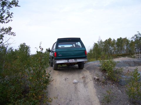 1996 Ford Bronco - A 1996 Ford Bronco navigates an off-road trail, showing its rear profile against a backdrop of trees and an overcast sky.
