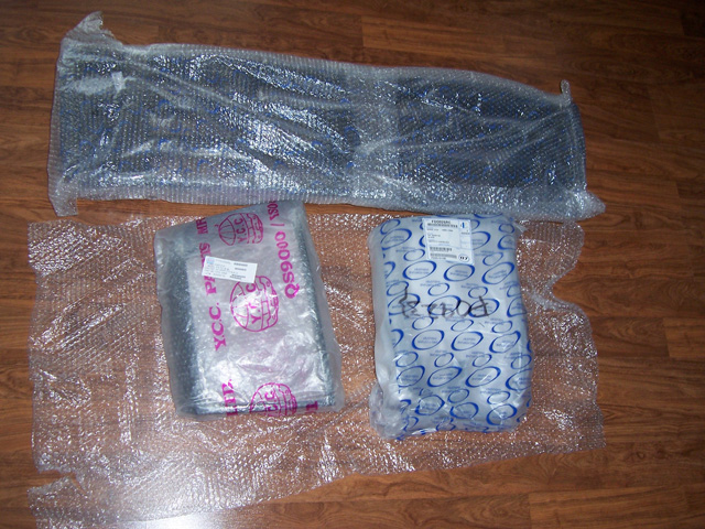 Three unopened auto parts in bubble wrap and plastic packaging laid out on a wood floor