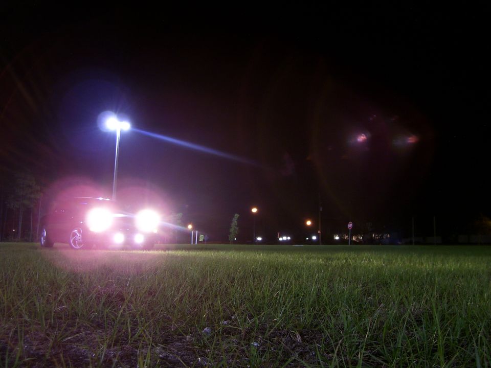 Car at night with headlights and fog lights on, shot low from a grassy field