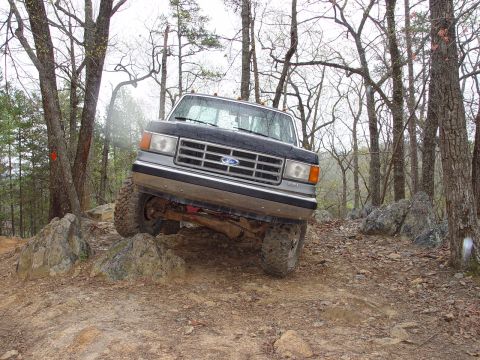 1996 Ford Bronco - A black Ford F-Series truck is shown off-roading on a dirt and rock path through a wooded area.
