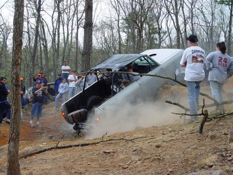 1996 Ford Bronco - An off-road vehicle is tilted on its side, kicking up dust as onlookers observe the scene in a wooded area.