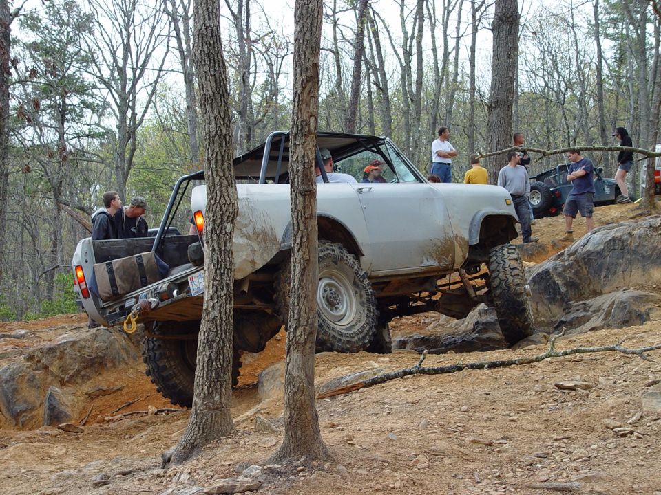 Off-road vehicle with roll cage climbing over rocks on a dirt trail, other vehicles and people in background