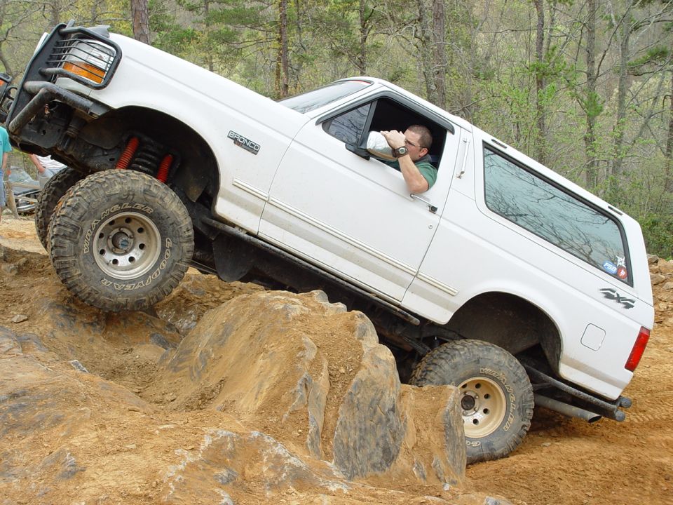 White 1996 Ford Bronco climbing over rocks on an off-road trail. Driver is holding a drink