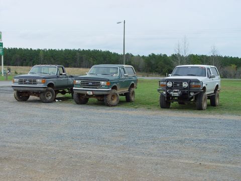 1996 Ford Bronco - Three lifted SUVs, including a green 1992 Ford Bronco, are parked in a gravel lot with a wooded background.