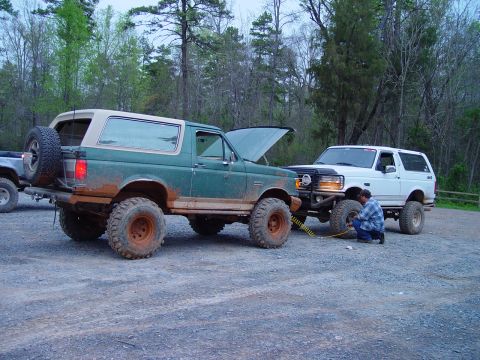 1996 Ford Bronco - Two lifted Ford Broncos are parked on a gravel lot, one with its hood up and a man working on a tire.