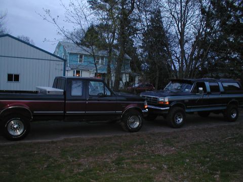 1979 Ford Bronco - Two pickup trucks are parked next to each other in a yard with houses and trees in the background.