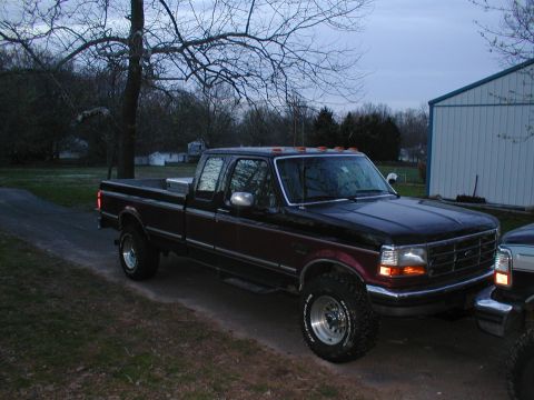 1979 Ford Bronco - A dark red and black Ford F-Series truck is parked on a dirt road near a building, with another vehicle partially visible.