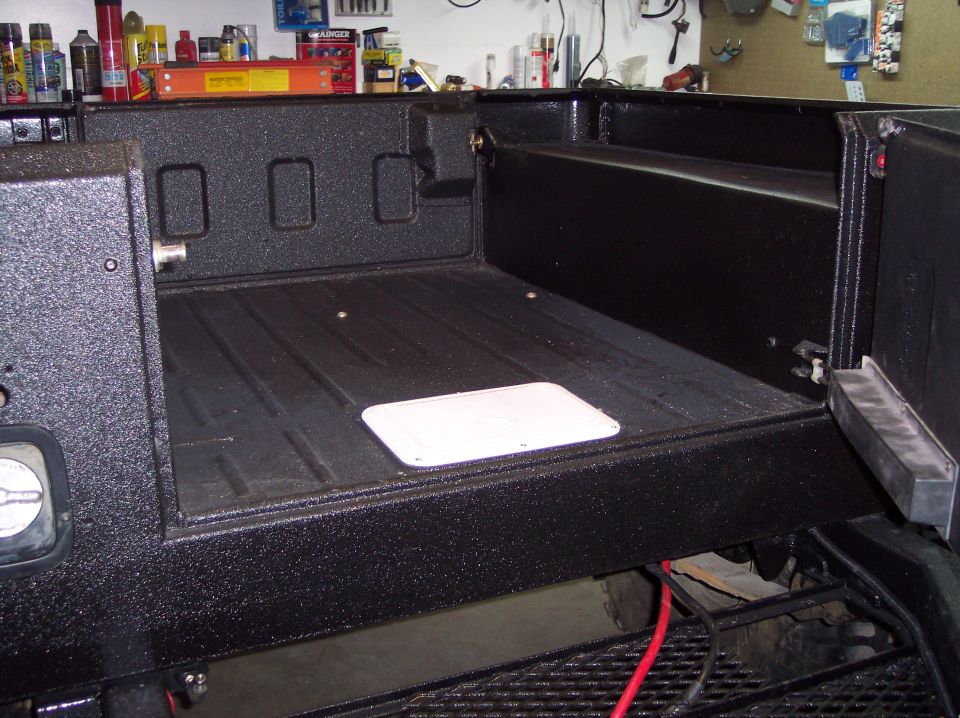 Bed of a vehicle with black textured liner, showing a white access panel