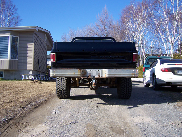 Rear view of lifted black square-body Chevy pickup with chrome bumper and mud tires on gravel