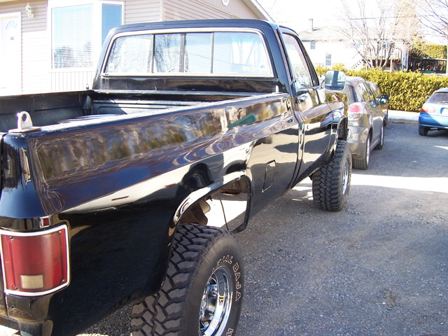 Black square-body Chevy pickup rear three-quarter view on chrome wheels and Baja mud tires