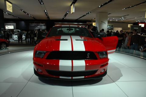 2005 Ford Mustang - A red Ford Mustang Shelby GT500 is on display at an auto show with people in the background.