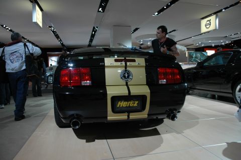 2005 Ford Mustang - A black Ford Mustang with gold racing stripes and a Hertz logo is displayed indoors, possibly at a car show.
