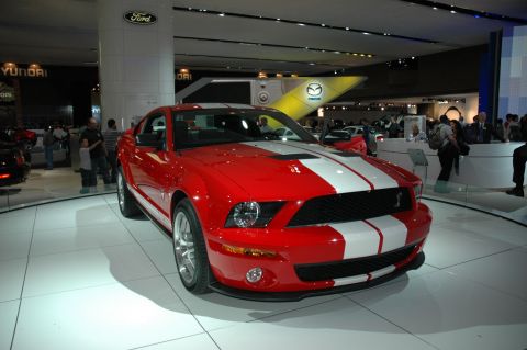 2005 Ford Mustang - A red 2005 Ford Mustang with white racing stripes is displayed at an auto show with other car brands visible in the background.