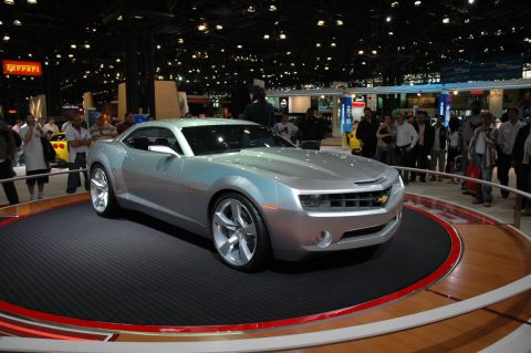 2005 Ford Mustang - A silver Chevrolet Camaro concept car is displayed on a rotating platform at an auto show with spectators in the background.