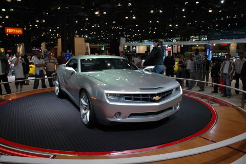 2005 Ford Mustang - A silver Chevrolet Camaro concept car is displayed on a rotating platform at an auto show, with spectators in the background.