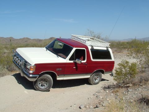 1990 Ford Bronco - Two-tone red and white 1990 Ford Bronco equipped with a roof rack sits on a dusty desert trail.